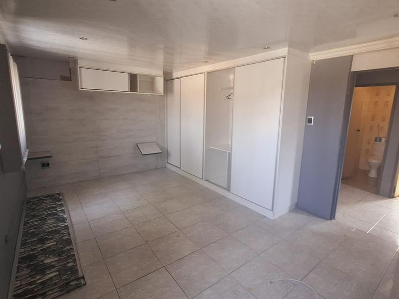 A spacious bedroom featuring tiled flooring, built-in wardrobes, and recessed ceiling lighting, with an adjacent bathroom visible through the doorway.