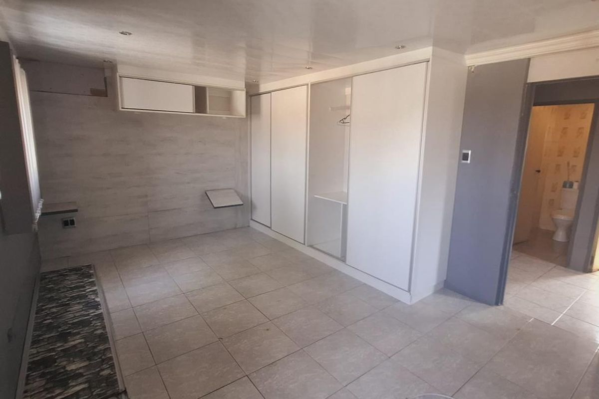 A spacious bedroom featuring tiled flooring, built-in wardrobes, and recessed ceiling lighting, with an adjacent bathroom visible through the doorway.