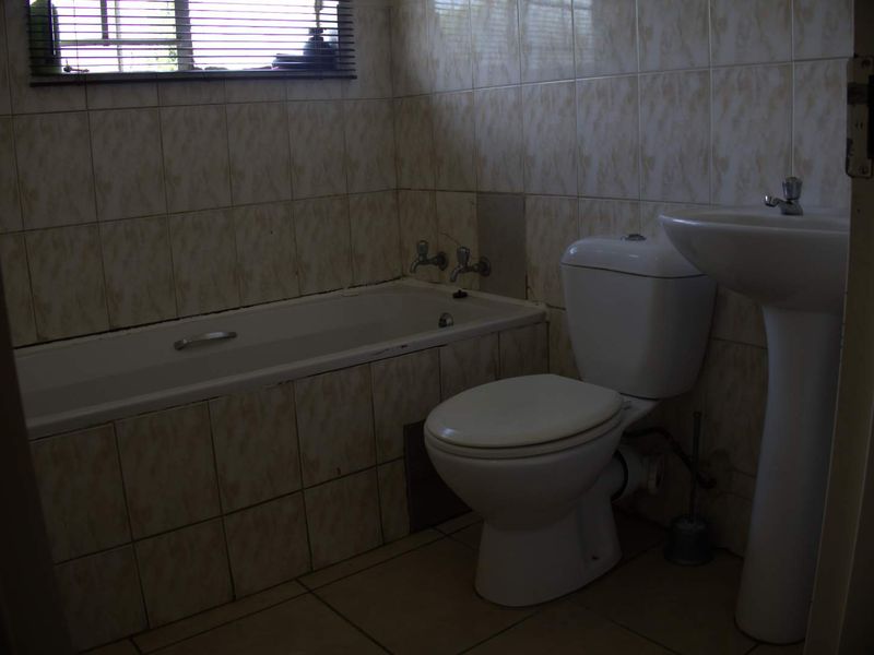A dated bathroom featuring a standard bathtub, pedestal basin, and toilet with full-height wall tiling and a window with a venetian blind.