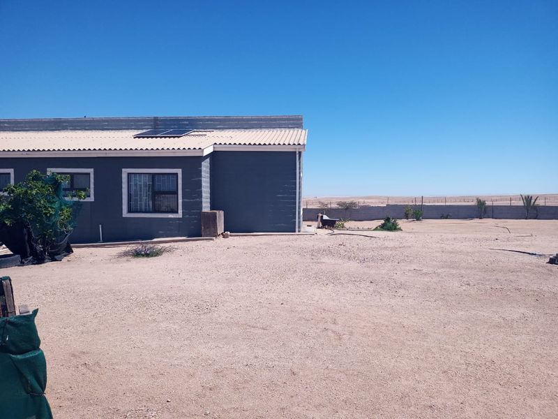 Rear view of the property showing the exterior wall, corrugated roof with solar panels, and a large open sandy yard area.