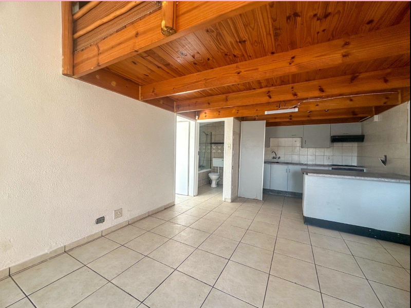 Open plan living area featuring exposed timber ceiling beams, tiled flooring, and a functional kitchen with breakfast counter, leading to an adjacent bathroom.