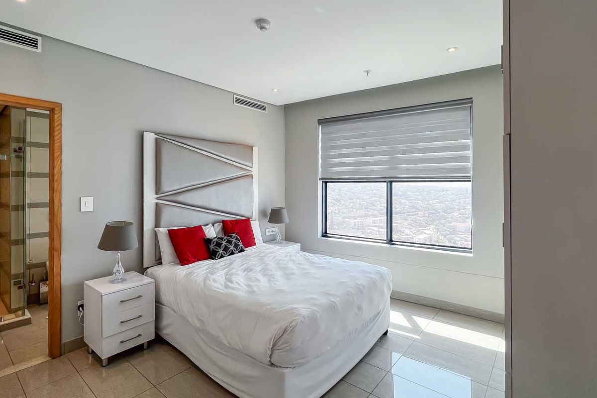 Modern bedroom featuring tiled flooring, ducted air conditioning, large window with roller blinds, and an en-suite bathroom with a glass-enclosed shower.
