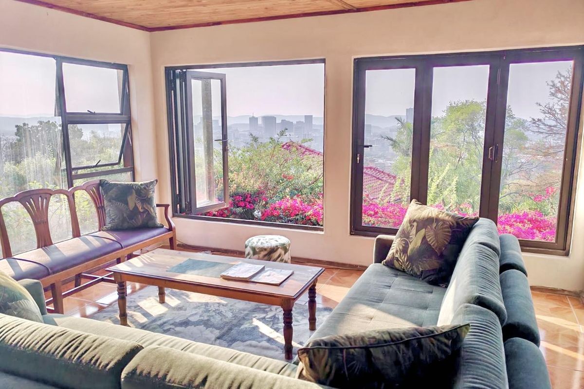 A bright living room featuring timber ceilings, tiled floors, and large aluminium windows offering expansive city views.