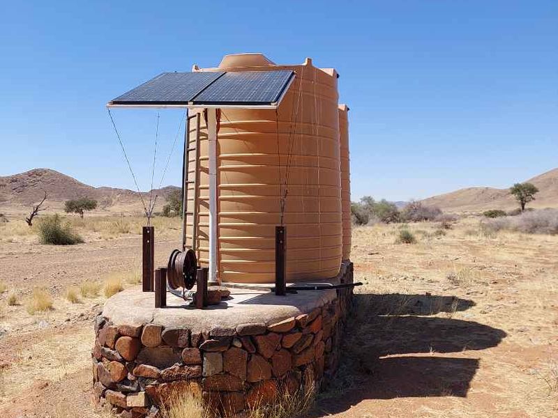 A solar-powered water storage system featuring large plastic tanks mounted on a stone-clad concrete base in a rural farm setting.