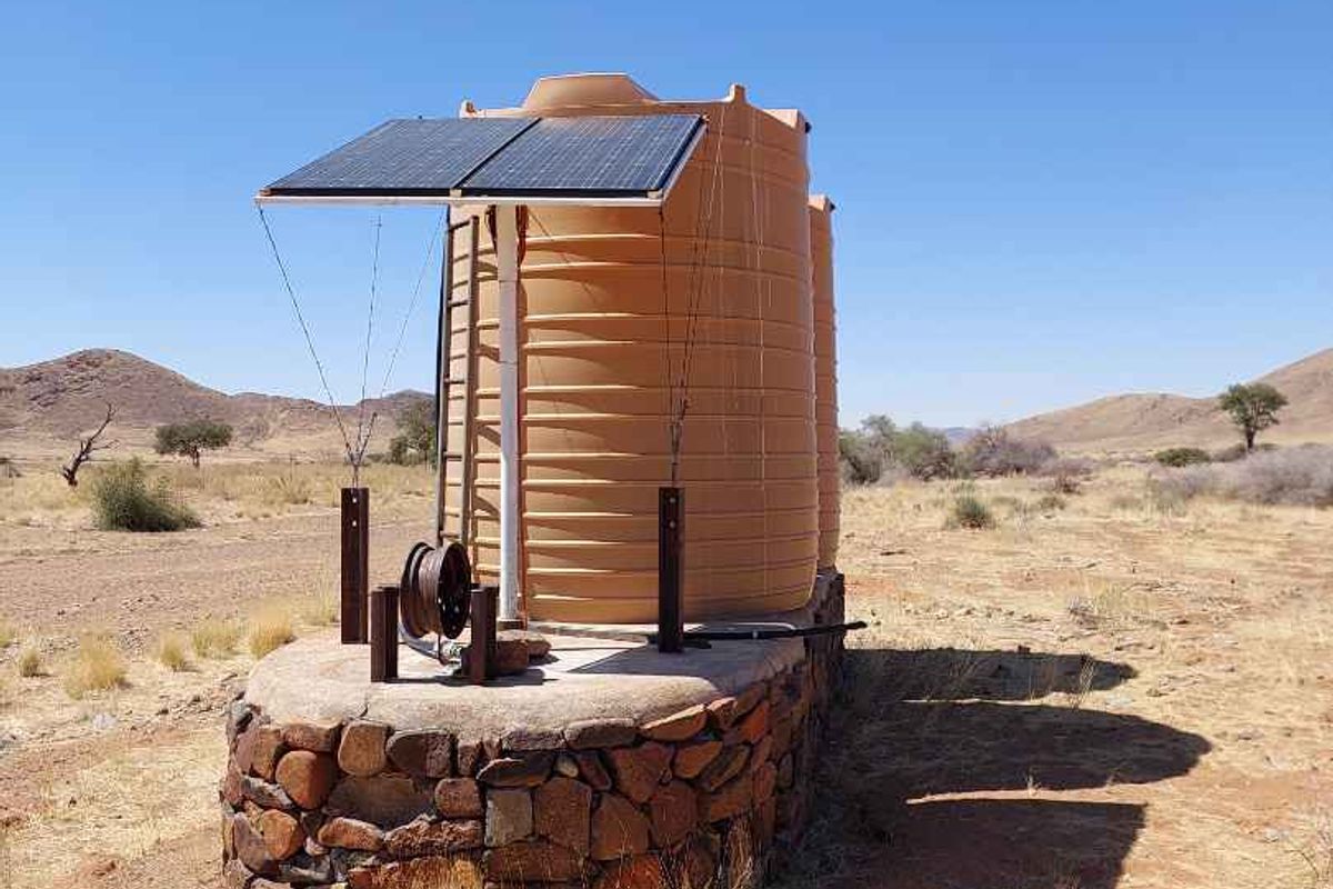 A solar-powered water storage system featuring large plastic tanks mounted on a stone-clad concrete base in a rural farm setting.