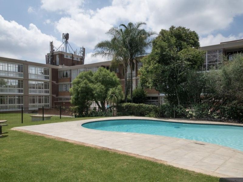 A well-maintained communal swimming pool area featuring a paved deck, landscaped garden, and metal perimeter fencing set against the apartment building facade.