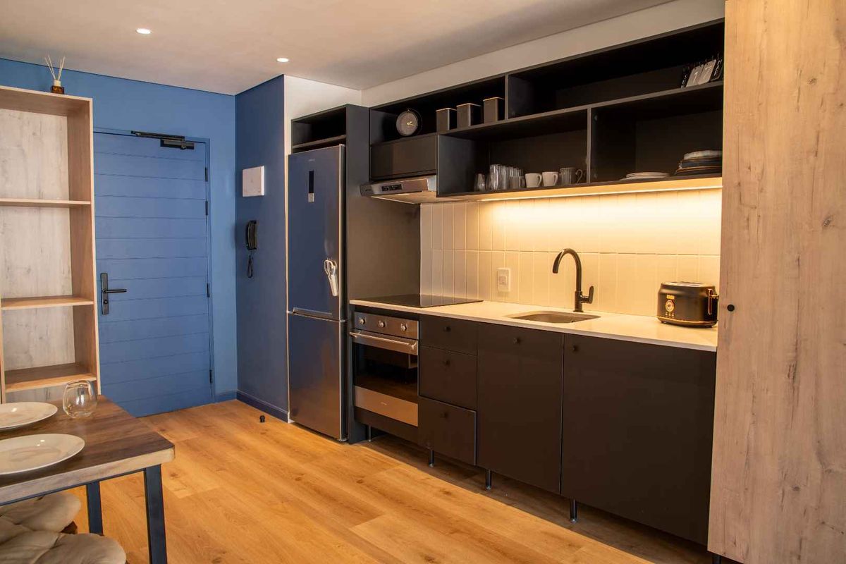 Modern compact kitchen featuring dark cabinetry, integrated oven, hob, extractor fan, and sleek under-cabinet lighting, set against a blue accent wall.