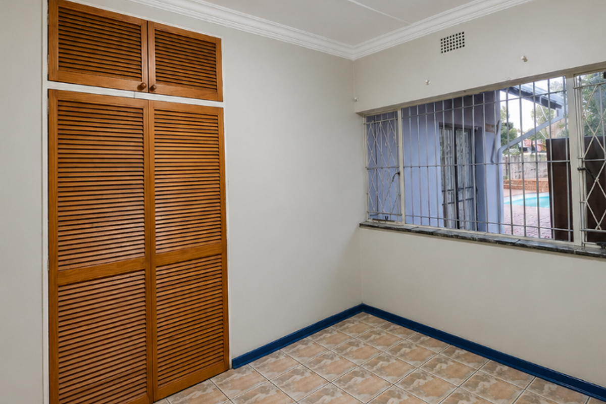 A bright, empty bedroom featuring tiled flooring, a built-in wooden louvered wardrobe, and a window fitted with security bars.