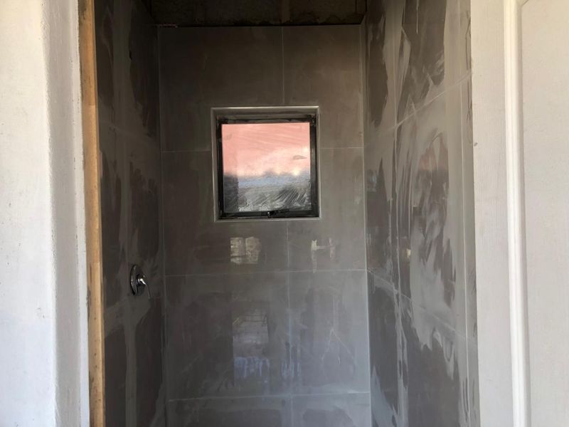 An unfinished bathroom shower enclosure featuring grey wall tiles, a shower mixer, and a small window, currently in the final stages of construction.