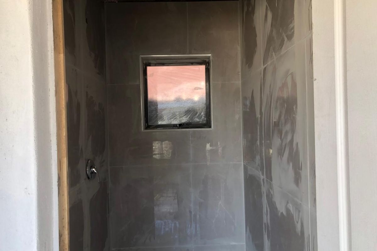 An unfinished bathroom shower enclosure featuring grey wall tiles, a shower mixer, and a small window, currently in the final stages of construction.