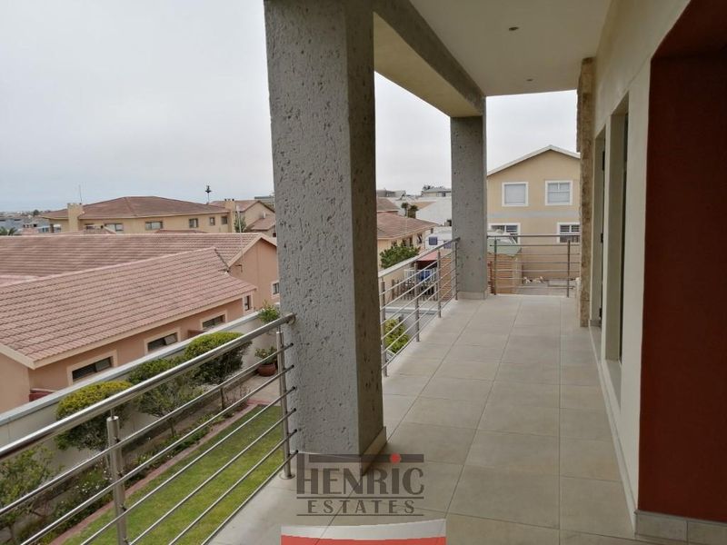 A spacious covered balcony featuring tiled flooring, stainless steel balustrades, and stone cladding, offering an elevated view of the surrounding residential area.