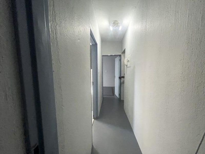 A simple, empty hallway with white painted walls and grey flooring, featuring a ceiling light and wall-mounted intercom unit.