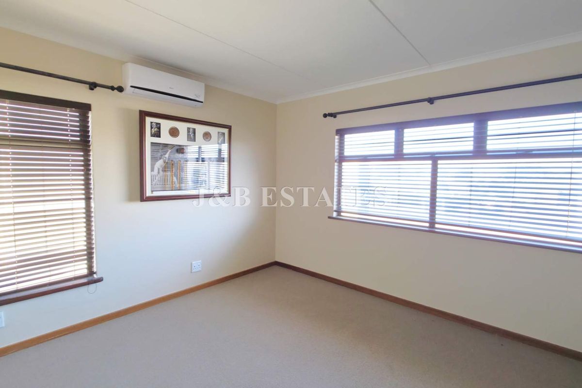 A bright, empty bedroom featuring neutral carpeting, wall-mounted air conditioning unit, and wooden venetian blinds on the windows.