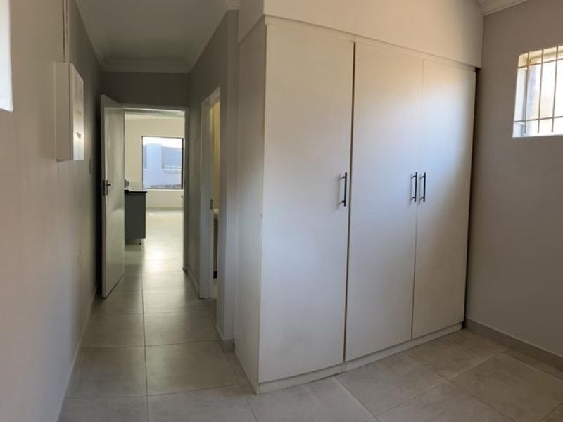 A clean, empty bedroom featuring neutral tiled flooring, built-in cupboards, and a window with security bars, leading into a hallway.
