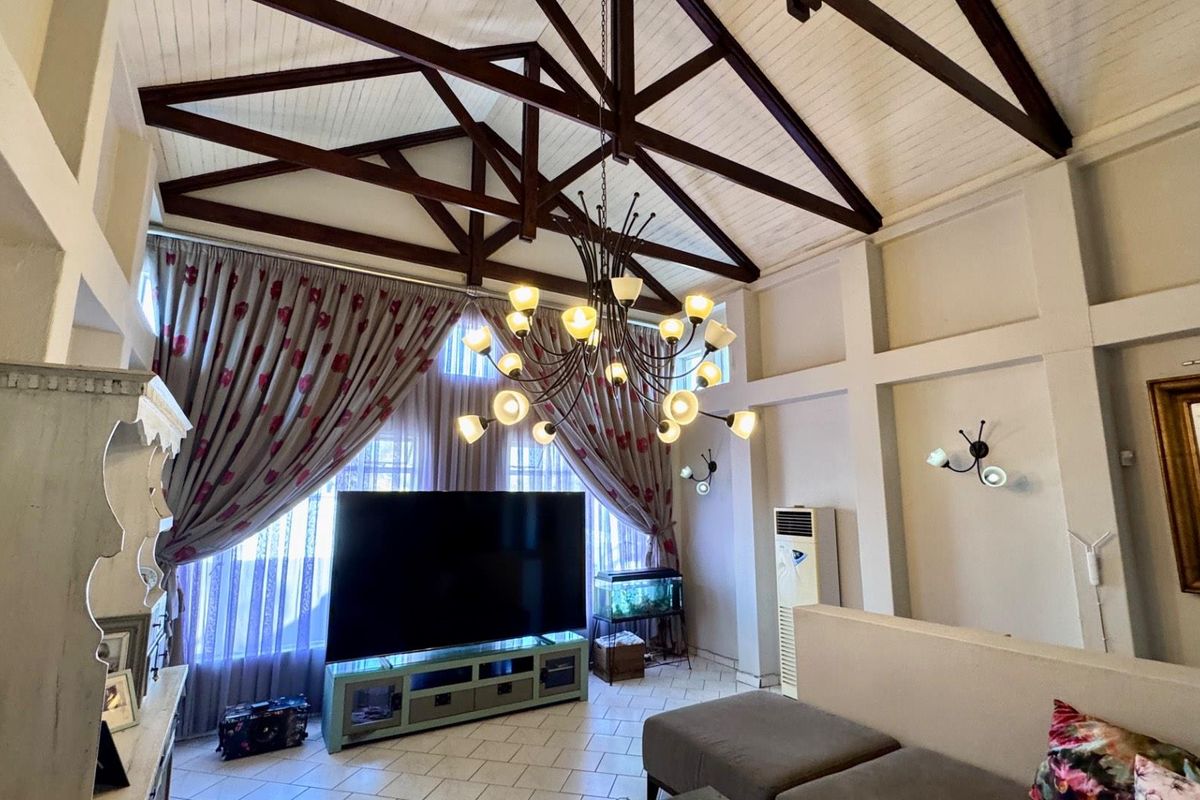 Spacious living room featuring high vaulted ceilings with exposed timber trusses, tiled flooring, and large windows with decorative curtains.