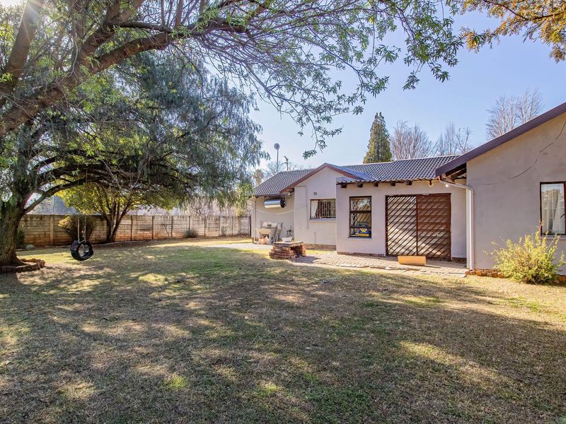 Spacious rear garden featuring a paved patio area, built-in braai, and established trees, enclosed by a secure brick boundary wall.