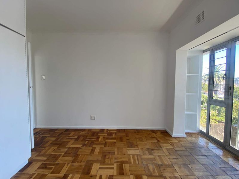 An empty bedroom featuring classic parquet flooring, built-in cupboards, and integrated shelving, with large windows providing natural light and exterior views.