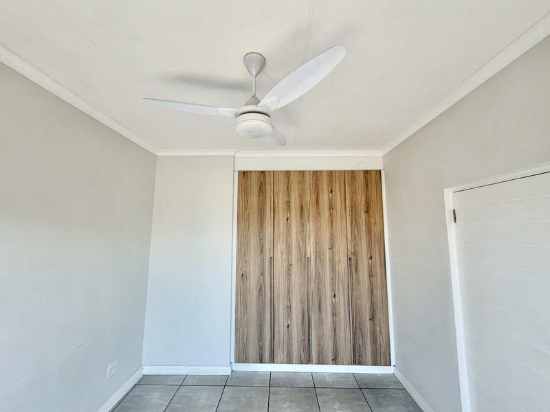 A clean, empty bedroom featuring modern wood-finish built-in cupboards, neutral wall paint, tiled flooring, and a central ceiling fan.