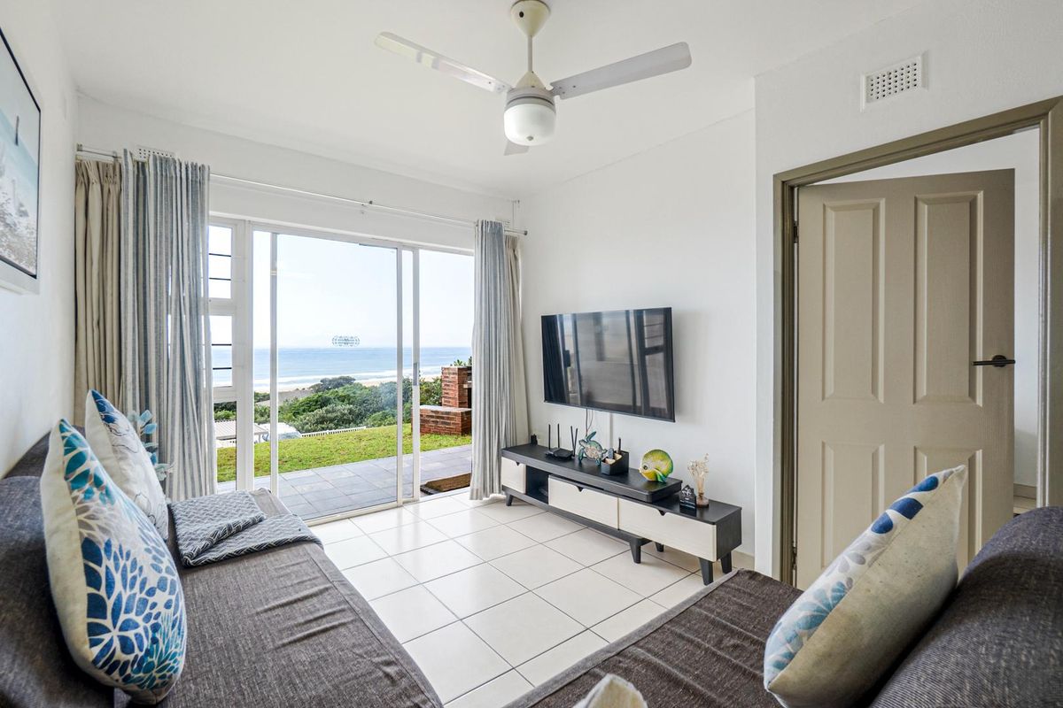 A bright living room featuring tiled flooring, a ceiling fan, and large sliding glass doors that open to a patio with ocean views.