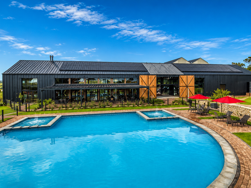 A modern industrial-style building featuring a large swimming pool with integrated spas, expansive paved deck, and scenic rural surroundings.