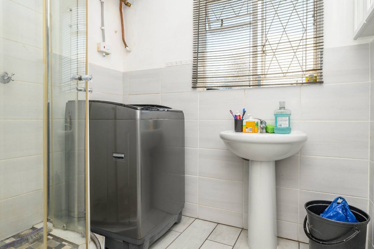 A functional bathroom featuring a pedestal basin, a shower enclosure, and space for a washing machine, finished with neutral wall and floor tiling.