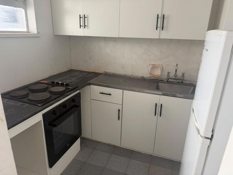 Compact kitchen featuring built-in oven, electric hob, stainless steel sink, and white cabinetry. The space requires cosmetic updates to wall finishes and backsplash.