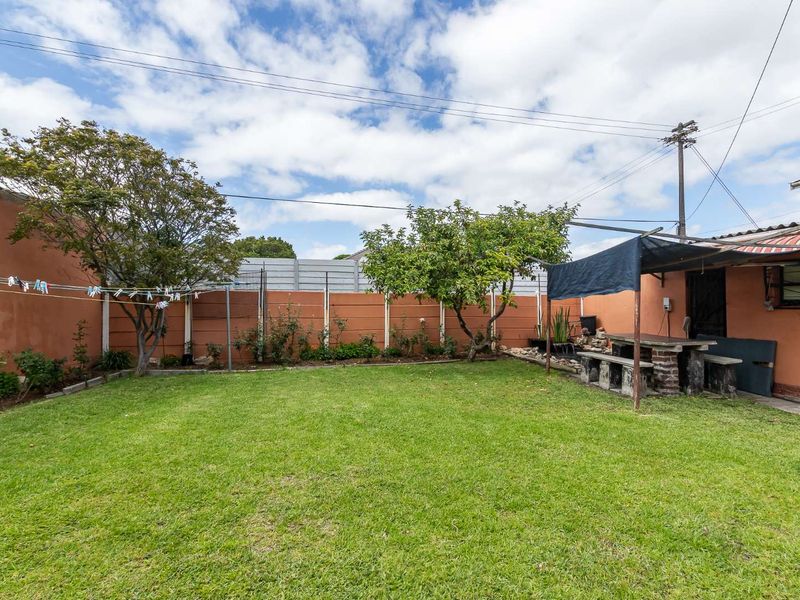 A private rear garden featuring a well-maintained lawn, perimeter boundary wall, and a built-in stone seating area under a covered structure.