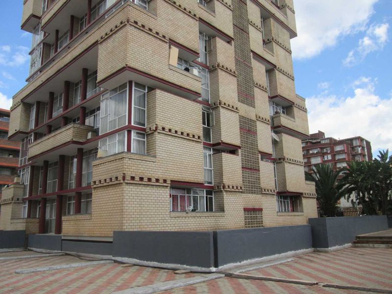Exterior view of a multi-story brick apartment building featuring a paved driveway and concrete boundary wall.