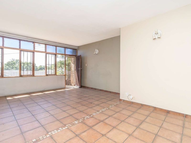 An empty, sunlit living area featuring tiled flooring, large windows with security bars, and neutral wall finishes, offering a blank canvas for renovation.