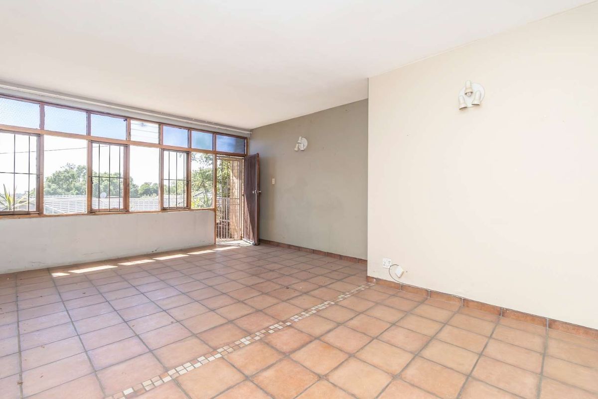 An empty, sunlit living area featuring tiled flooring, large windows with security bars, and neutral wall finishes, offering a blank canvas for renovation.