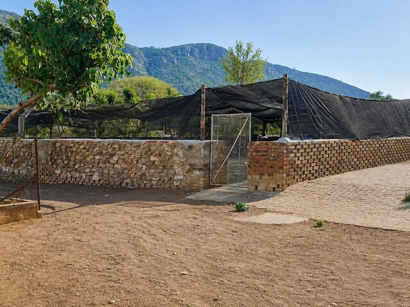 An outdoor agricultural or nursery area featuring a brick and stone boundary wall, shade netting structure, and a metal gate with mountain views.