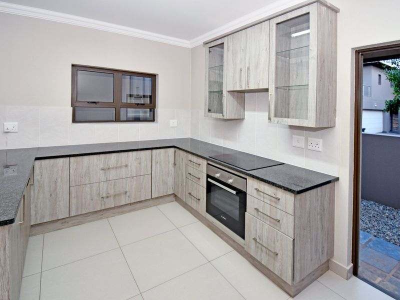 Modern kitchen featuring light wood-grain cabinetry, granite countertops, built-in oven, electric hob, and tiled flooring with direct access to an outdoor area.