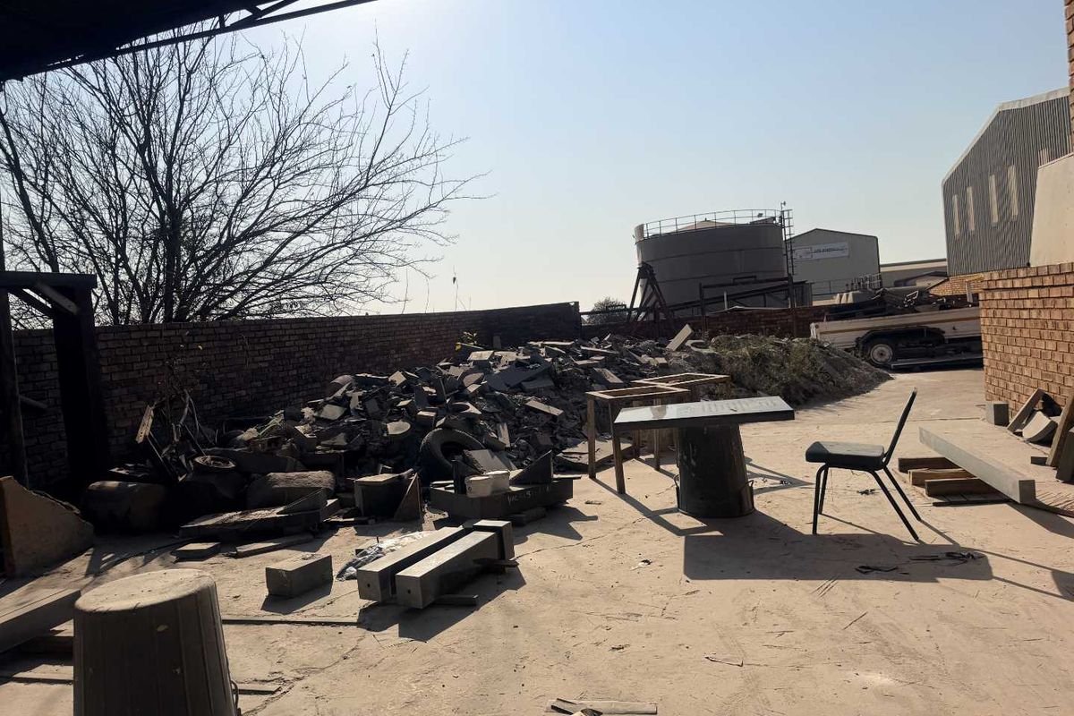 An industrial rear yard area featuring a concrete surface, brick boundary wall, and metal roofing structure, currently used for storage and site maintenance.