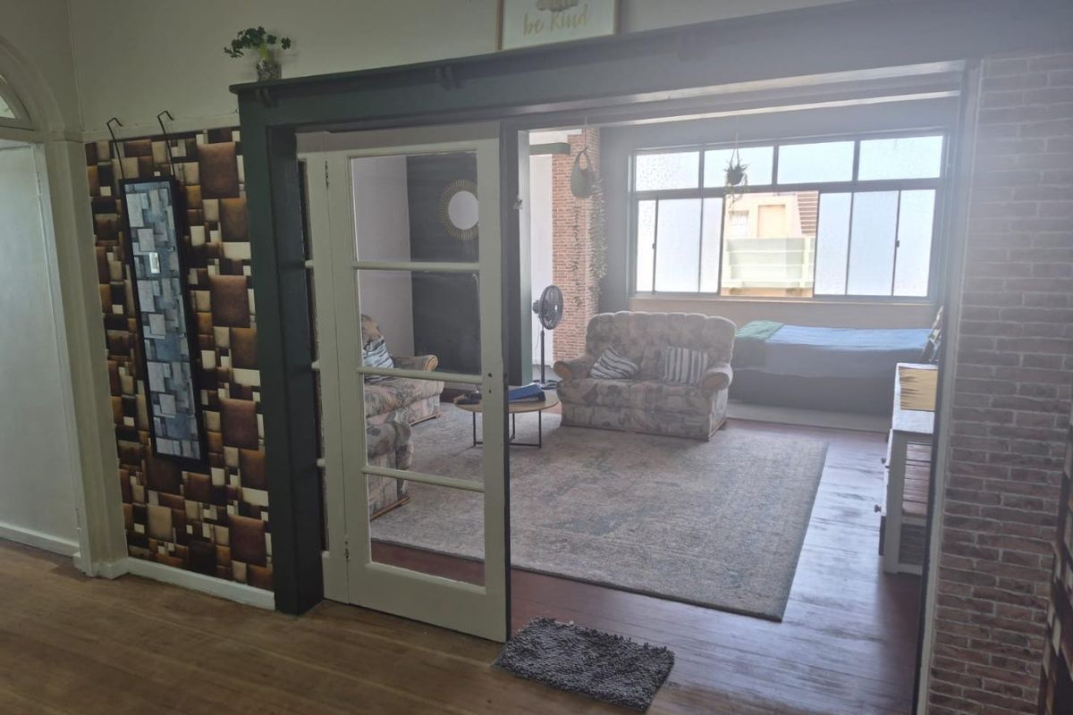 A living room featuring timber flooring, an exposed brick wall, and classic French doors leading to an adjacent space with large windows.
