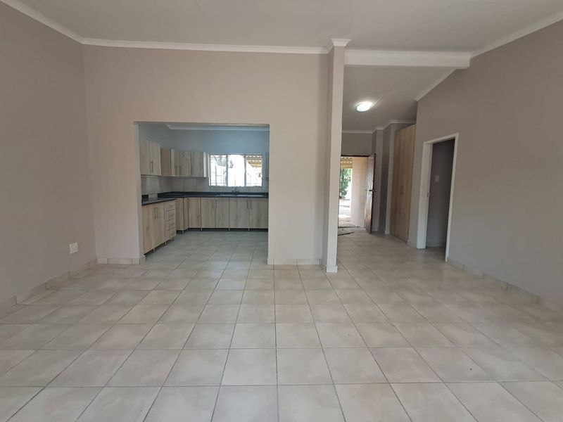 An open-plan living area featuring neutral tiled flooring, coved cornicing, and a view into a functional kitchen with built-in cabinetry and laminate countertops.