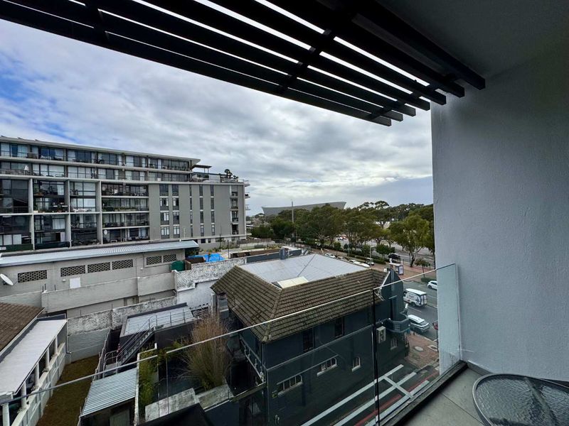 A modern apartment balcony featuring a glass balustrade and a slatted overhead pergola, offering an elevated urban view.