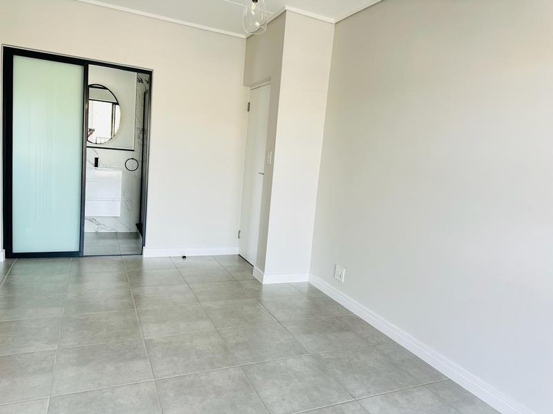 A bright, empty bedroom featuring neutral walls, modern grey floor tiles, and a sliding door leading to an en-suite bathroom.