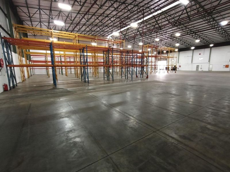 Spacious industrial warehouse featuring high ceilings, exposed steel trusses, high bay lighting, and extensive built-in racking systems on polished concrete flooring.
