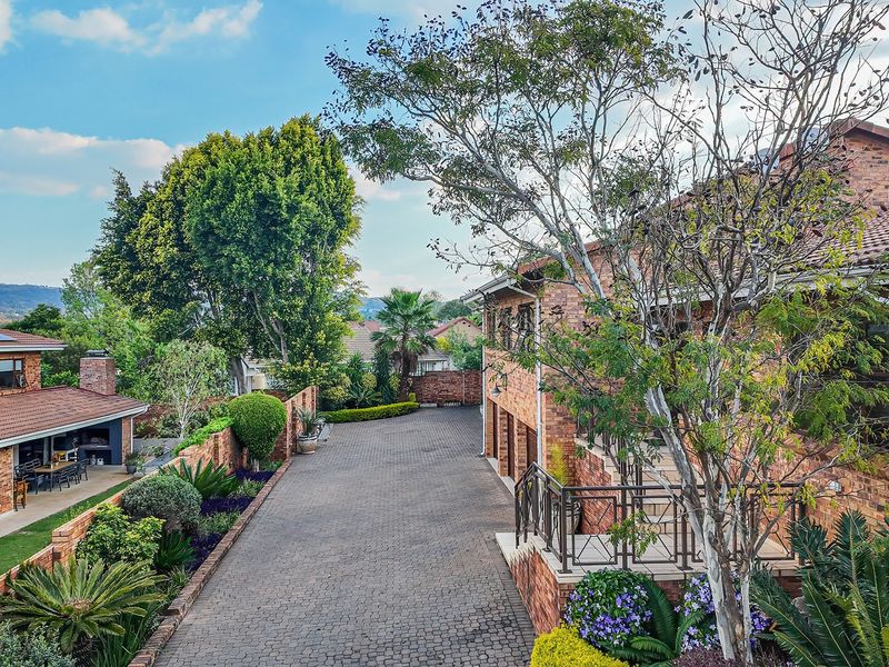 An elevated view of a brick-faced residential property featuring a spacious paved driveway, landscaped garden areas, and a covered patio with a built-in fireplace.