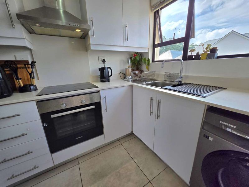 A compact, well-maintained kitchen featuring white cabinetry, a built-in oven, electric hob, extractor fan, and a stainless steel sink with a window view.
