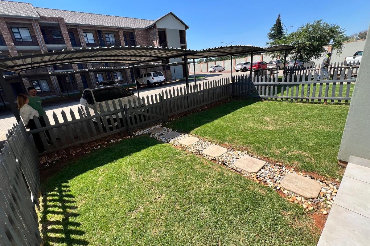 A private rear garden area featuring a manicured lawn, stone pathway, and perimeter timber fencing, adjacent to the townhouse complex parking area.