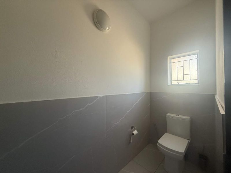 A clean and functional bathroom featuring modern grey wall tiles, a white toilet, and a small window with security bars.