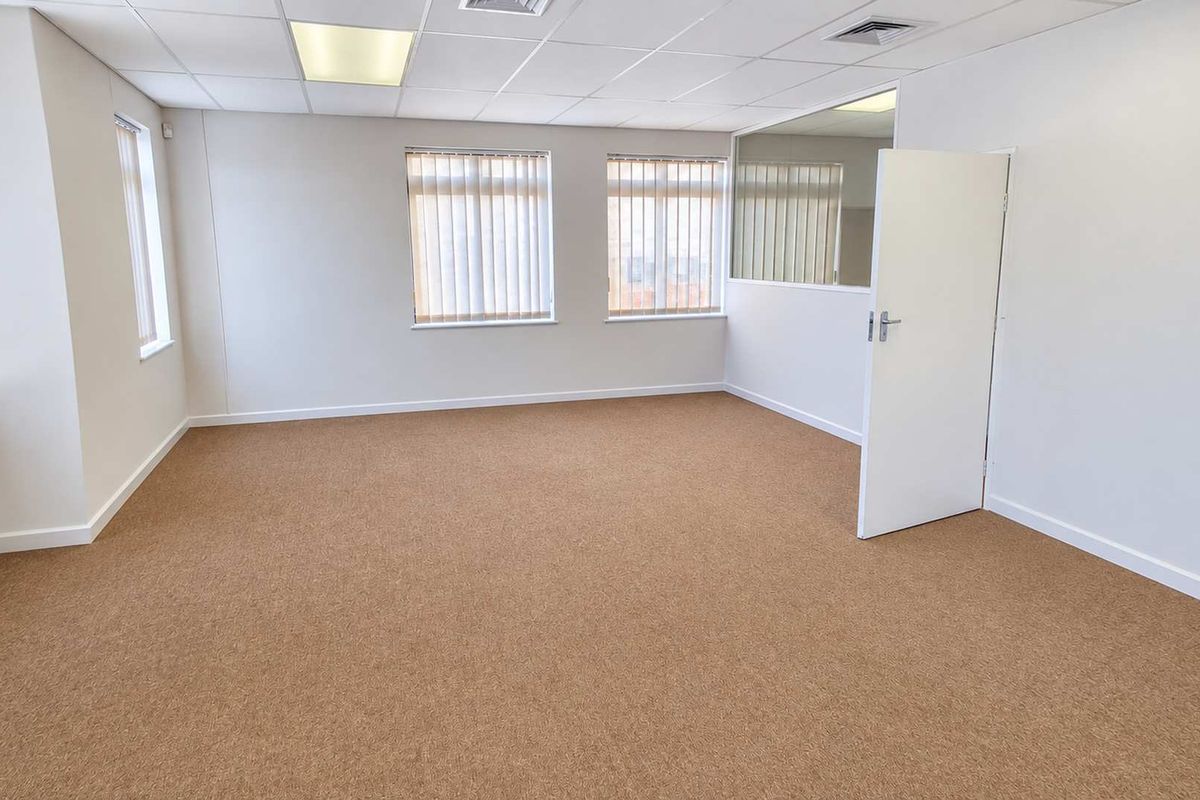 A bright, vacant office space featuring neutral carpet flooring, a suspended ceiling with recessed lighting, and large windows with vertical blinds.