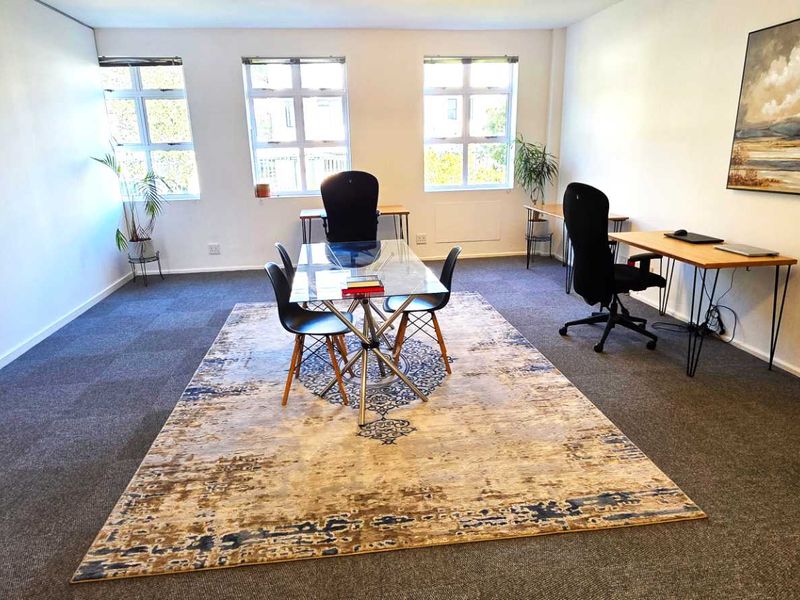 A functional office space featuring carpeted flooring and large windows that provide natural light, suitable for a professional work environment.