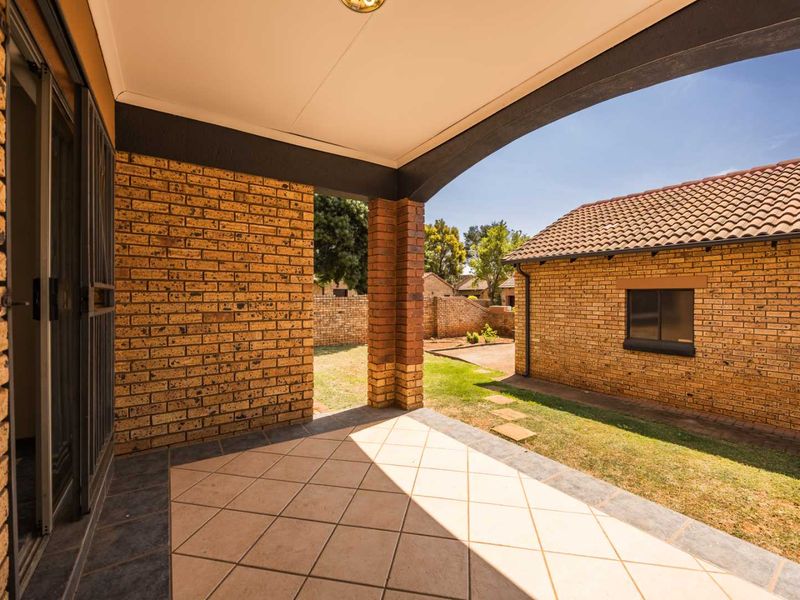 A covered patio area with tiled flooring and brick pillars, overlooking a private garden and adjacent brick structure.