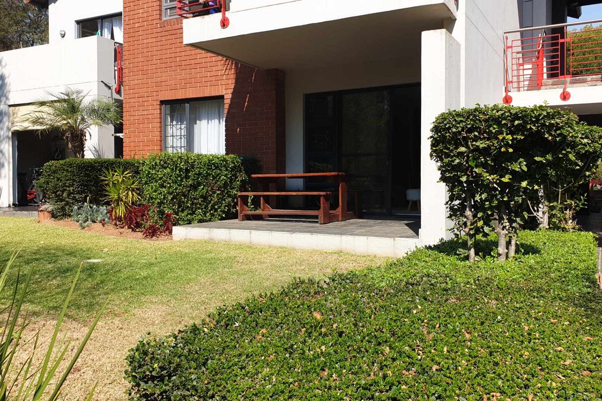 A ground-floor apartment featuring a private tiled patio, brick exterior detailing, and direct access to a well-maintained communal garden area.