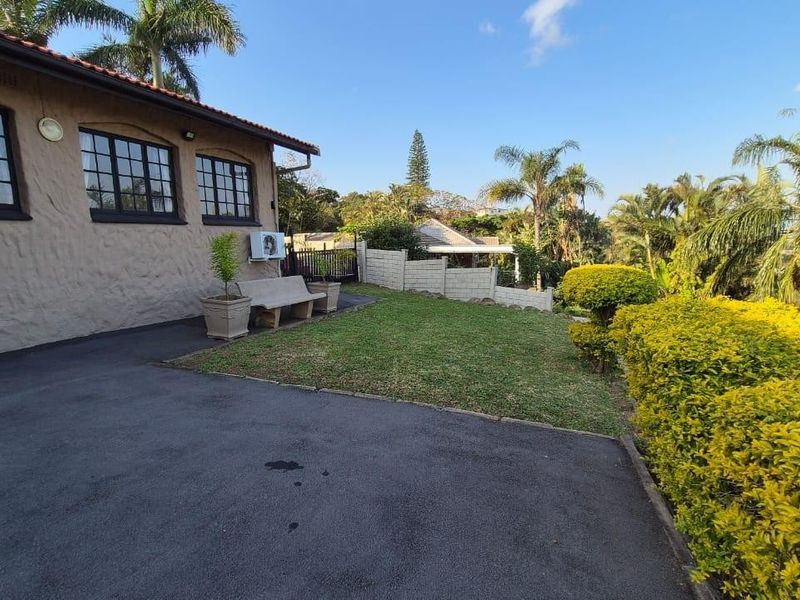 A well-maintained rear exterior view featuring a paved driveway, landscaped garden, boundary wall, and a house with a tiled roof and air conditioning unit.