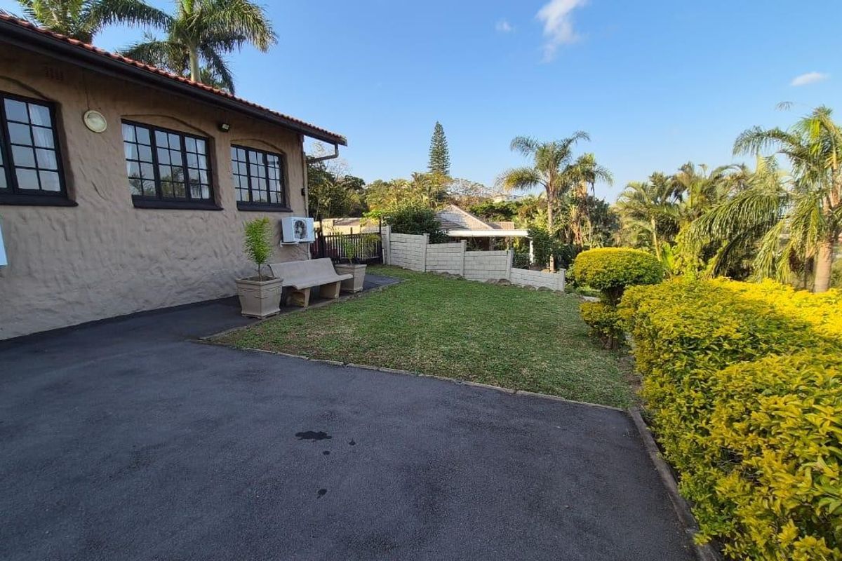 A well-maintained rear exterior view featuring a paved driveway, landscaped garden, boundary wall, and a house with a tiled roof and air conditioning unit.