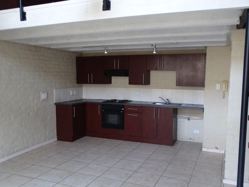 A compact kitchen featuring dark wood cabinetry, built-in oven and hob, tiled flooring, and an exposed brick wall, situated beneath a mezzanine level.