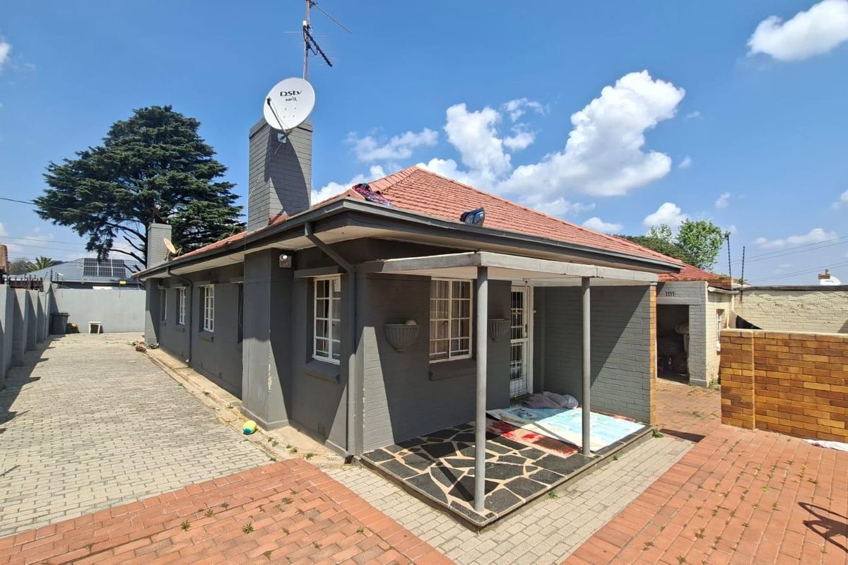Rear view of a single-story house featuring a covered patio, paved driveway, brick chimney, and secure perimeter walling with electric fencing.